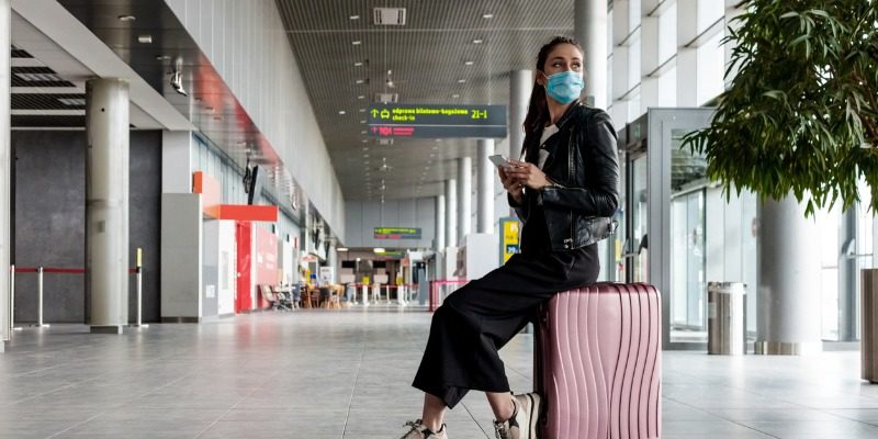 woman at the airport sitting on luggage with face mask on